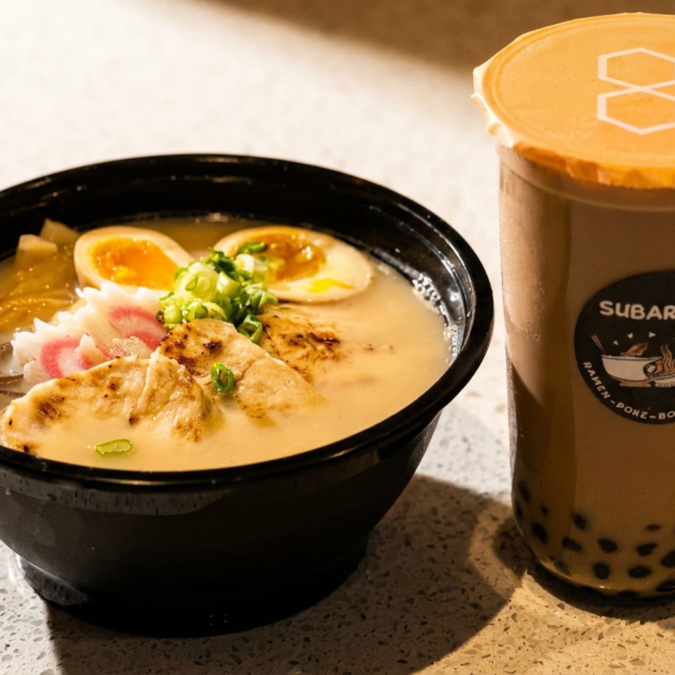 A bowl of ramen with chicken and eggs next to a Subarashi boba milk tea.