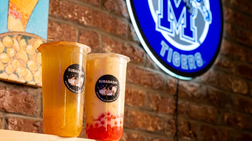 Two Subarashi bubble teas, one yellow and one creamy with red boba, against a brick wall and "M Tigers" sign.